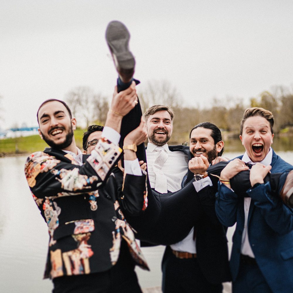Bräutigam wird von Freunden beim Gruppenfoto am Seeufer des Lago Ulm bei Hochzeit hochgehoben.